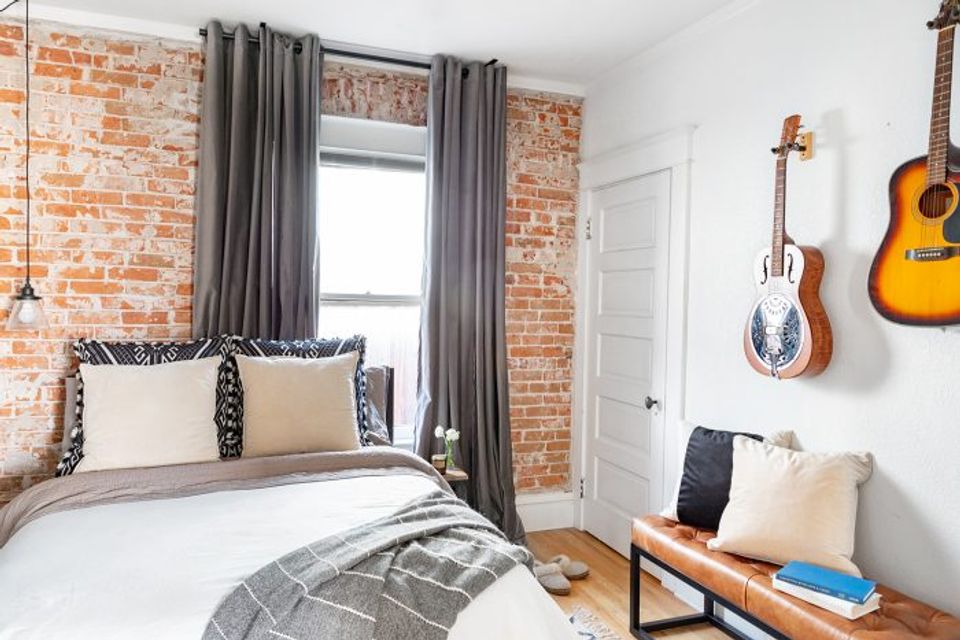 Eclectic bedroom with exposed brick accent wall, guitars as wall art, charcoal curtains, and bohemian touches.