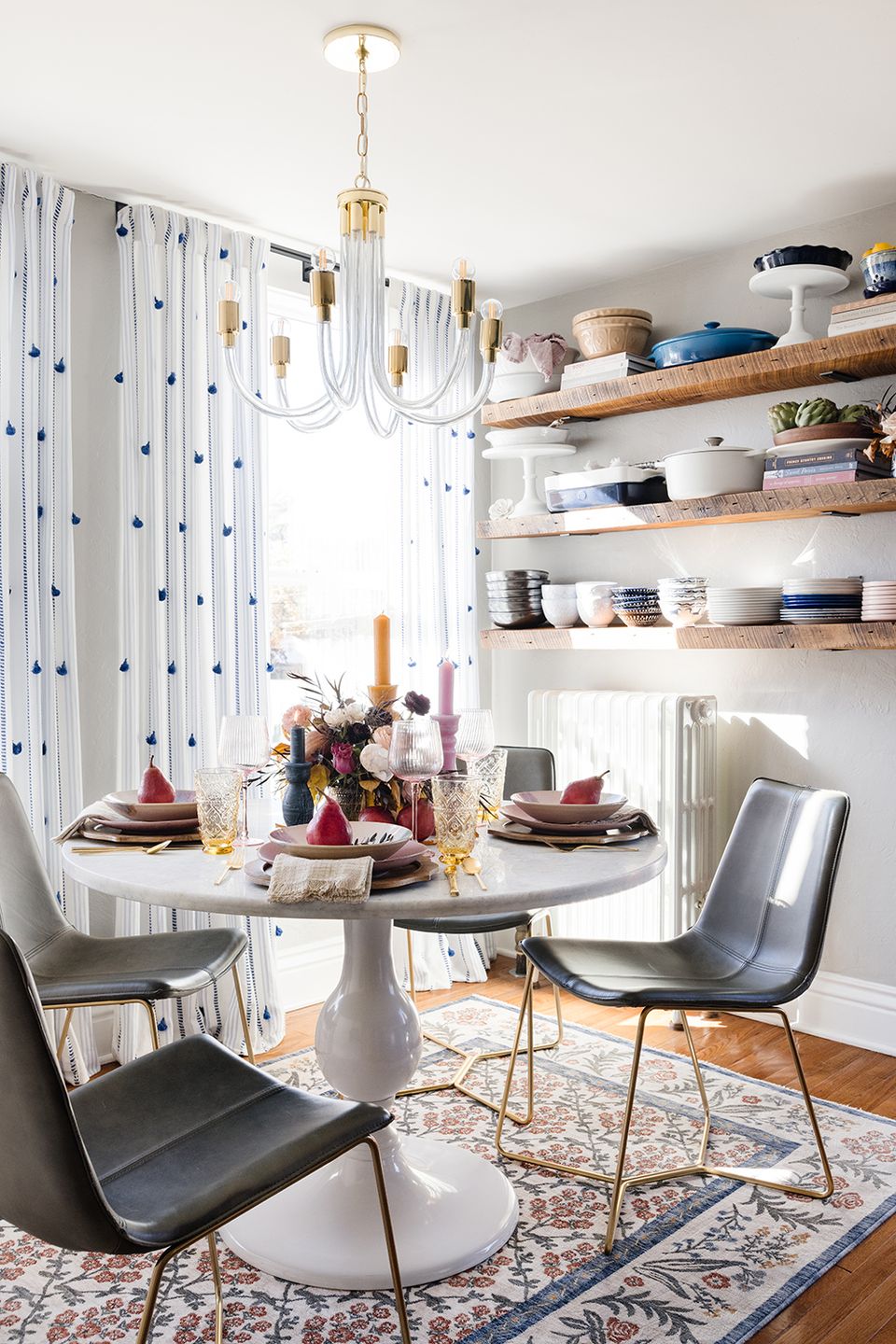 Contemporary dining nook with floating wood shelves, round pedestal table, patterned curtains, and brass chandelier