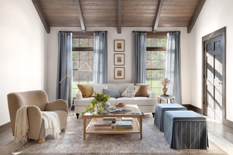 Rustic farmhouse seating area with light upholstery, layered pillows, wood beams, and airy blue drapery