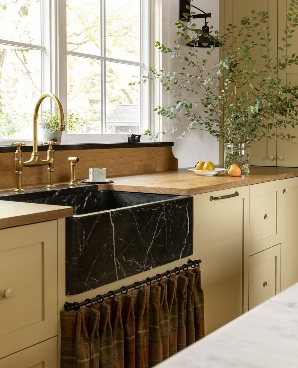 Warm sage-green kitchen with black marble sink, brass fixtures, and natural light from large windows creating an earthy, botanical aesthetic