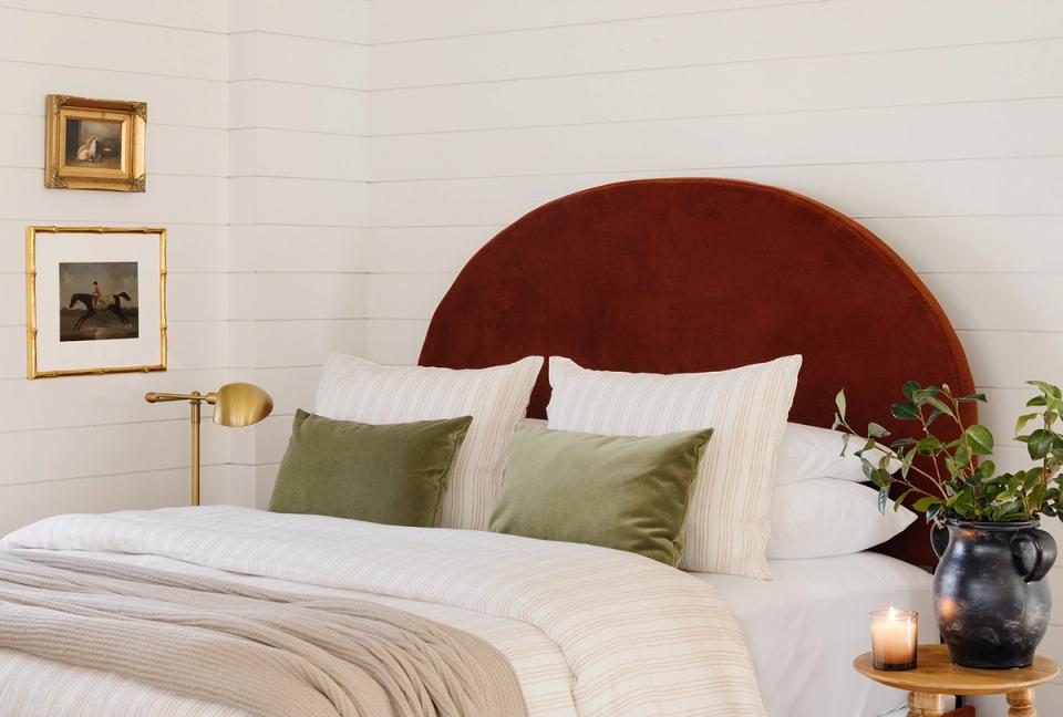 Bedroom with burgundy velvet curved headboard, sage green pillows, cream bedding, and vintage framed artwork