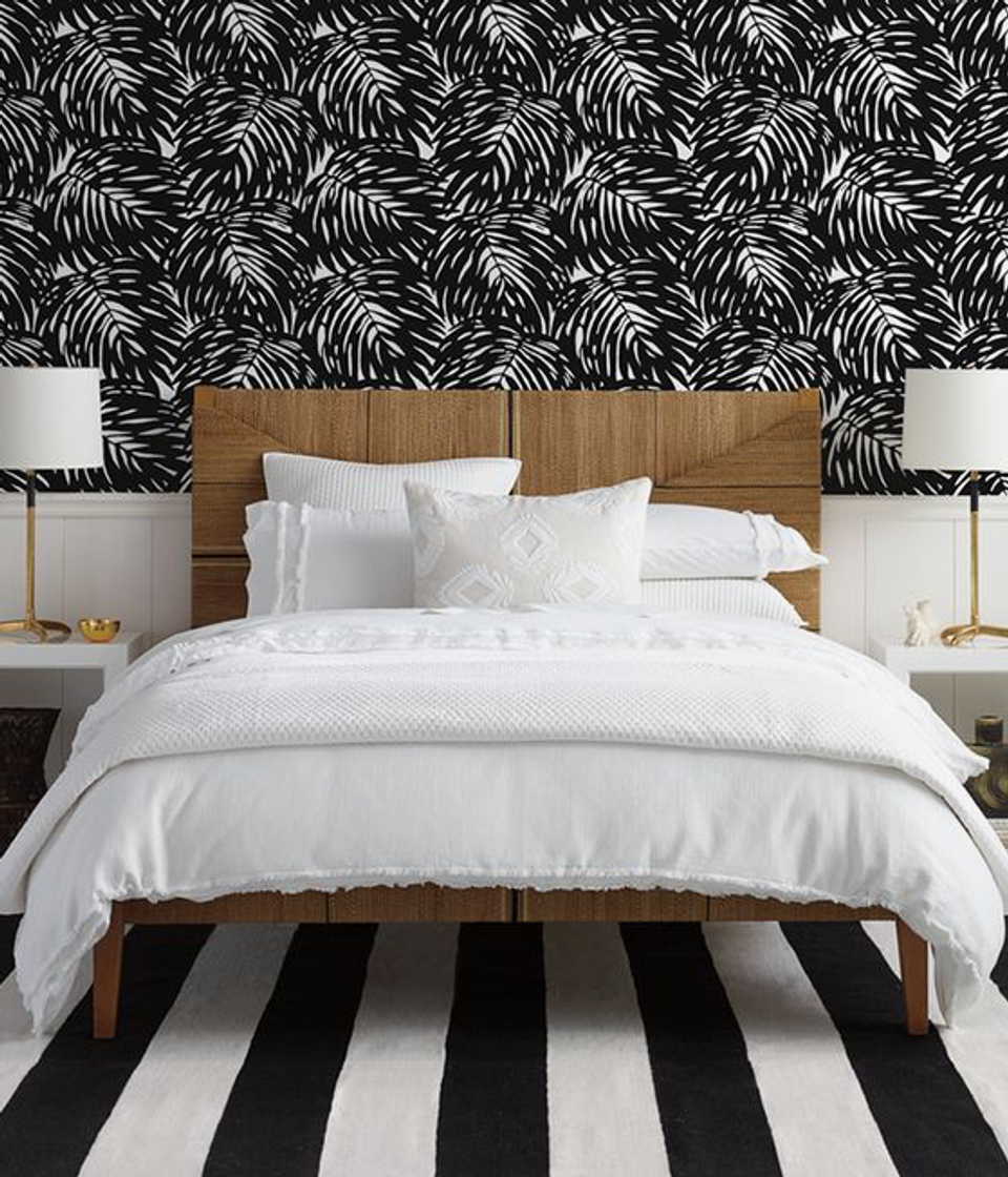 Modern bedroom with bold black palm-print wallpaper, warm wood headboard, crisp white bedding, and graphic striped area rug
