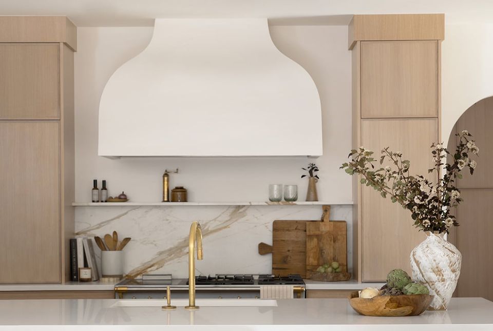 Minimalist kitchen with warm wood cabinetry, marble backsplash, and sculptural white hood in neutral palette