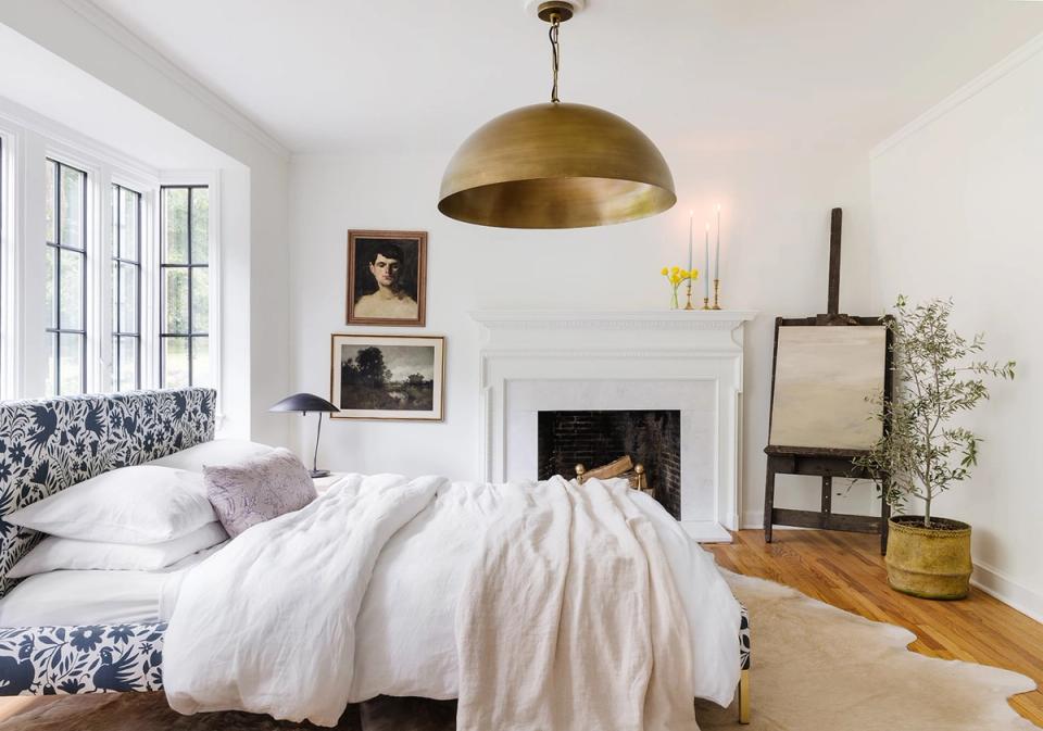 Eclectic modern bedroom with white fireplace focal point, brass pendant light, patterned blue headboard, and mixed vintage-industrial style elements.