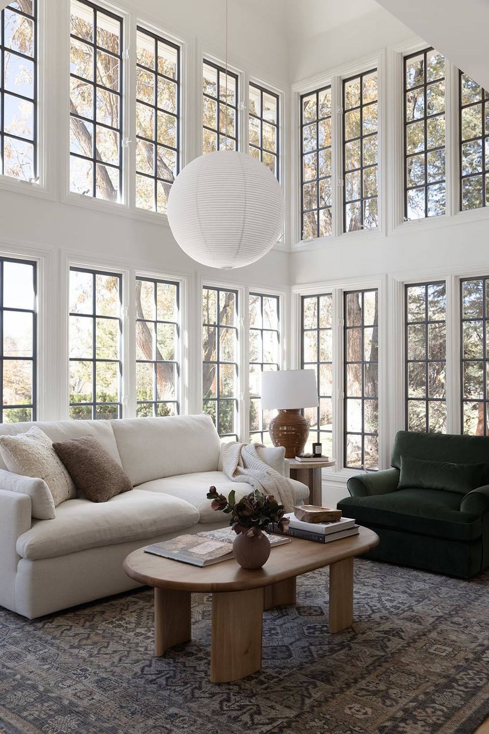 Bright organic modern living room with soaring windows, cream seating, natural wood base table, and airy sculptural pendant light