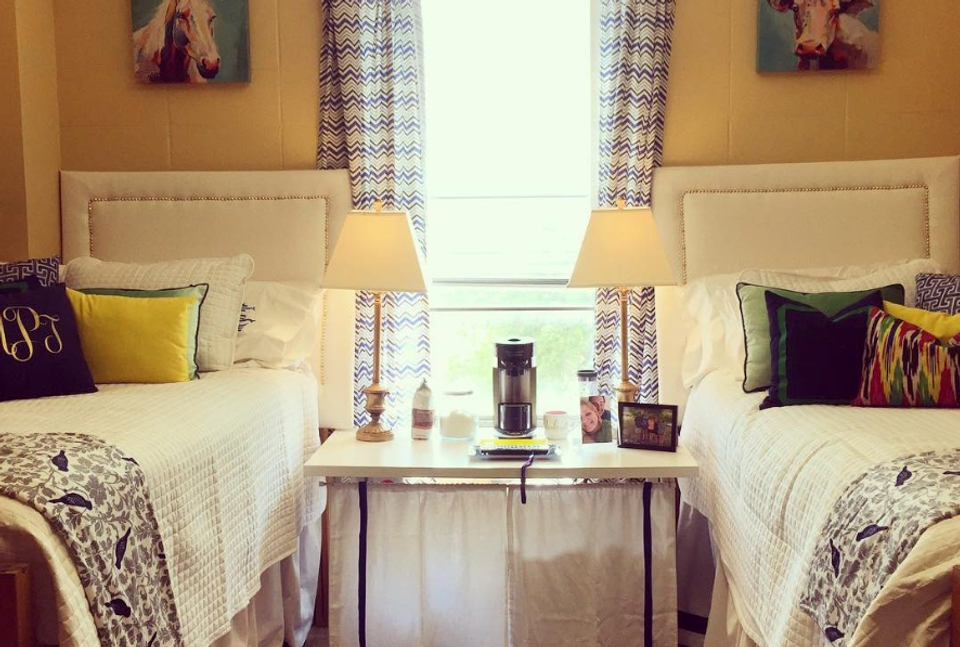 Warm, preppy dorm room with two twin beds, mustard walls, patterned curtains, and coordinated bedding in cream and navy tones