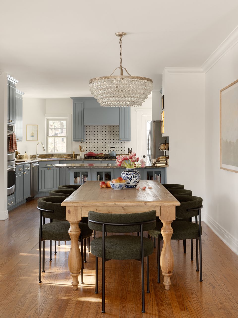 Refined transitional kitchen with soft blue cabinetry, natural wood table, black chairs, and crystal light