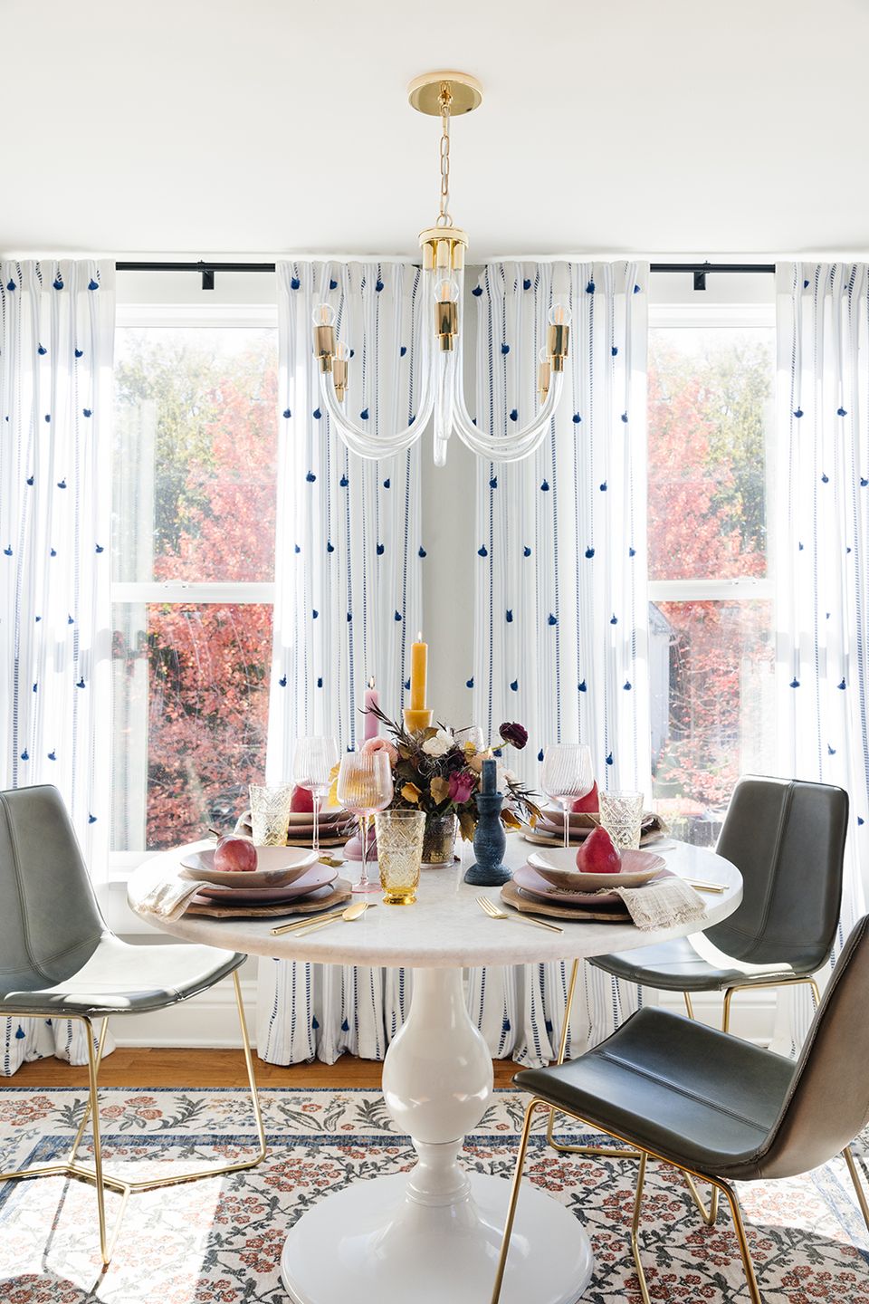 Elegant transitional round dining room with brass chandelier, patterned curtains, and modern chrome and gray chairs