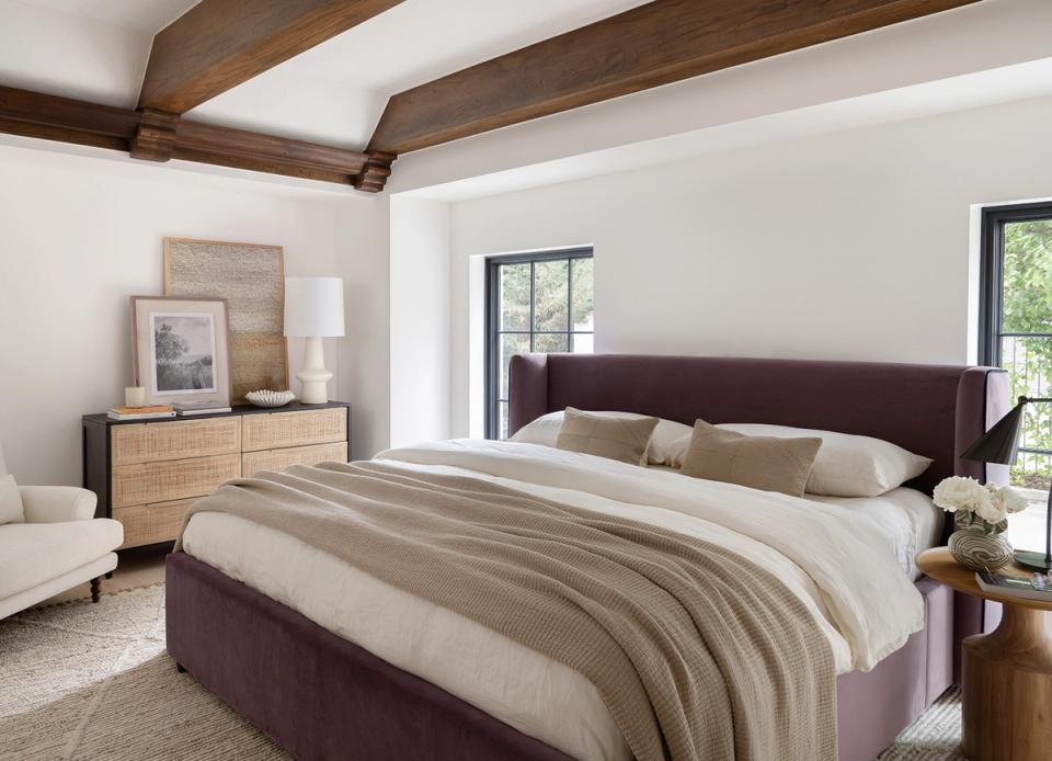 Modern farmhouse bedroom with dark purple storage bed, warm wood beams, neutral bedding, and natural light