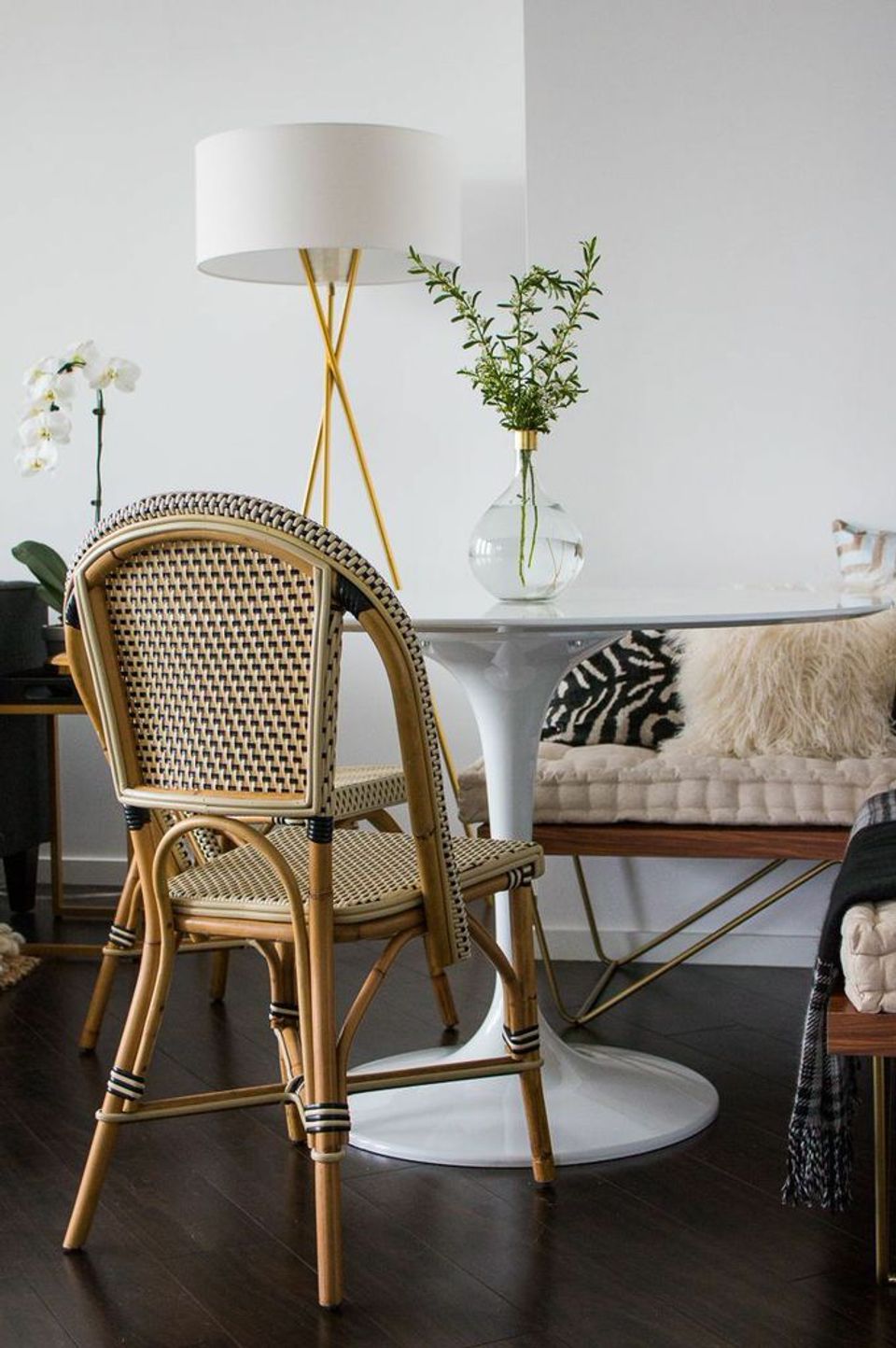 Modern French bistro corner with woven rattan chair, brass tripod lamp, and potted greenery on dark wood flooring.