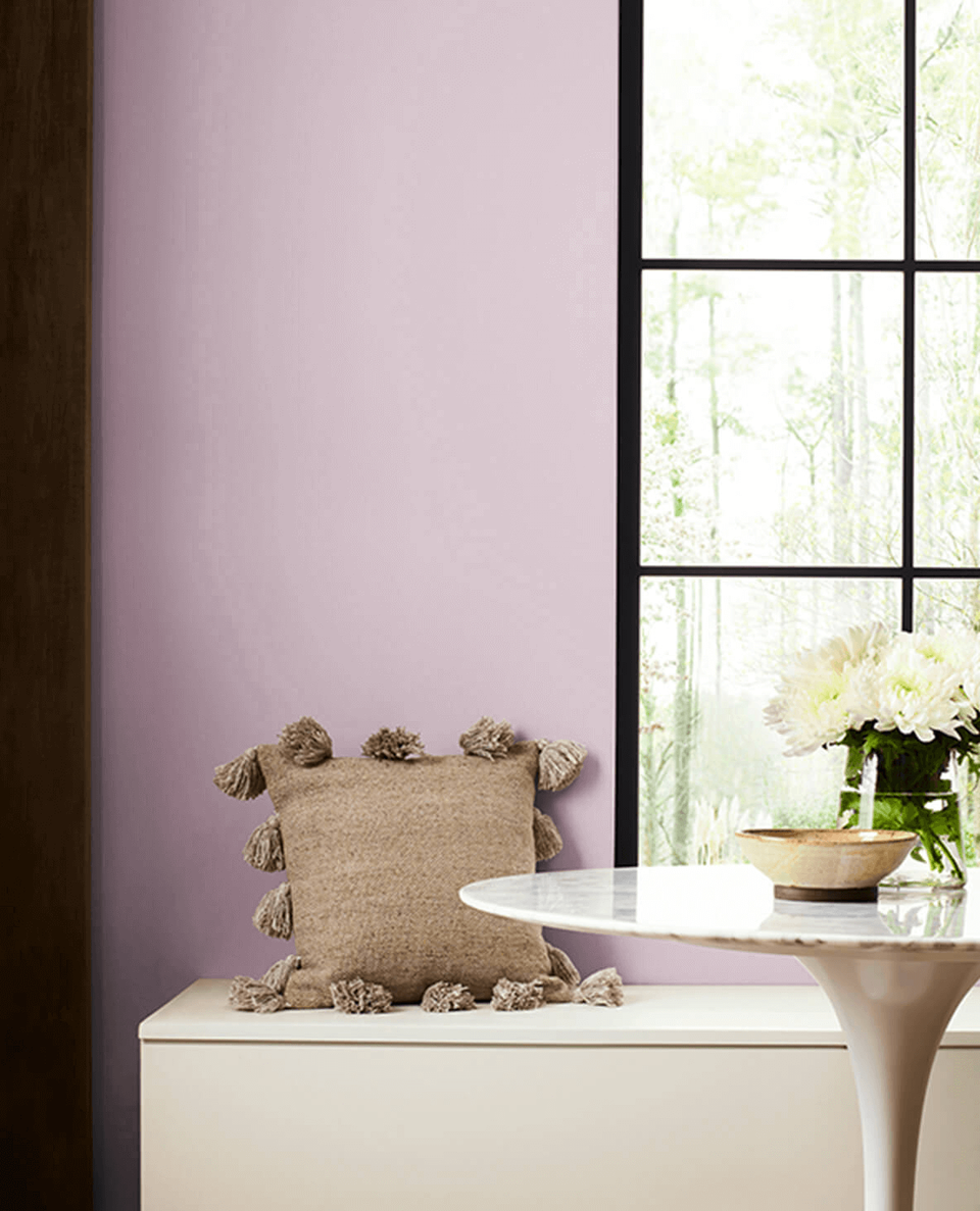 Mauve wall with tassel-trimmed pillow on white pedestal beside floor-to-ceiling black-frame windows and fresh flowers