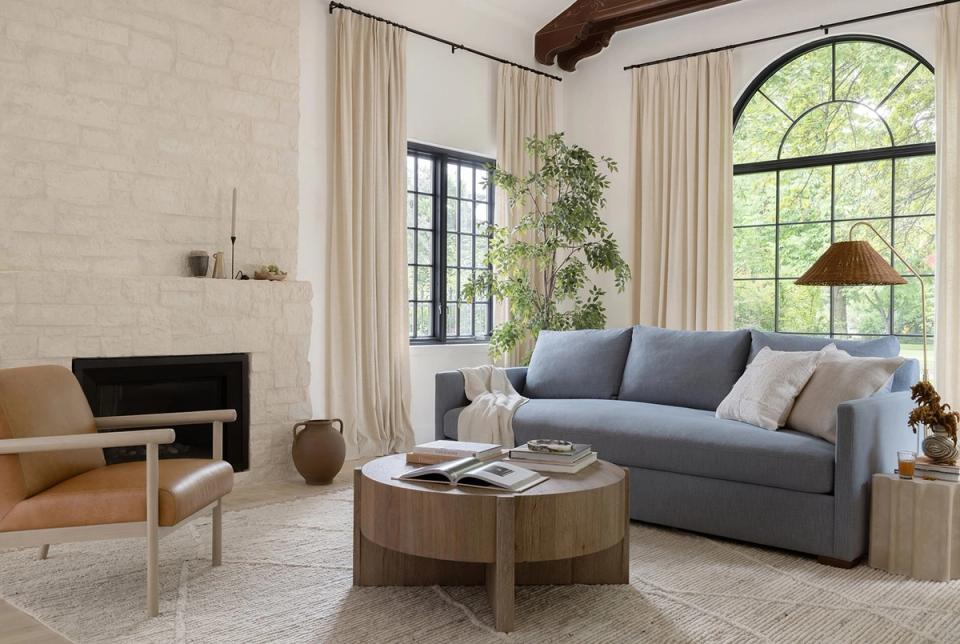 Modern Scandinavian living room featuring a slate blue sofa with warm wood accents, cream linen curtains, and abundant natural light from arched windows