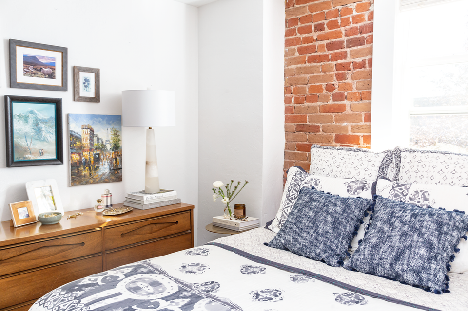 Eclectic bedroom with exposed brick wall, mid-century wood dresser, indigo patterned bedding, and gallery wall art.