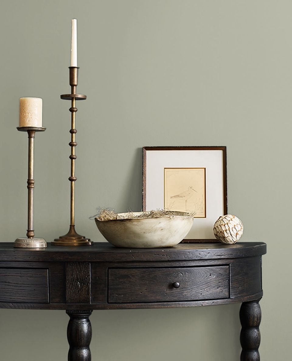 Eclectic vignette on dark console table against sage green wall with brass candlesticks, natural textures, and botanical artwork creating warm sophistication