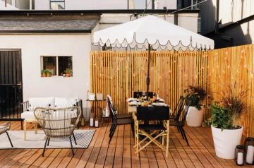 Warm mid-century modern patio with wooden deck, rattan chair, black dining table, and white scalloped awning against natural wood fence.