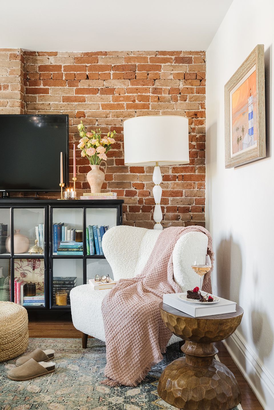 Eclectic reading corner blending industrial brick wall, cream chair, pink knit throw, and vintage brass side table with warm accents