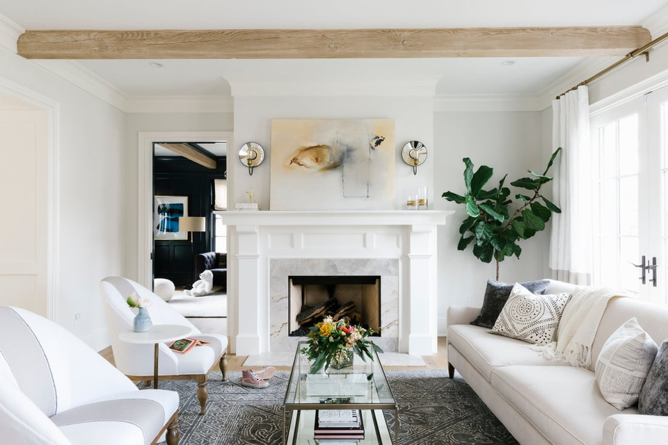Bright minimalist living room with white fireplace, neutral furnishings, natural wood beam, and lush greenery accents