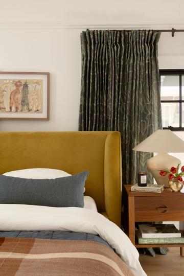 Modern traditional bedroom with mustard upholstered headboard, sage green textured curtains, and warm wood nightstand.