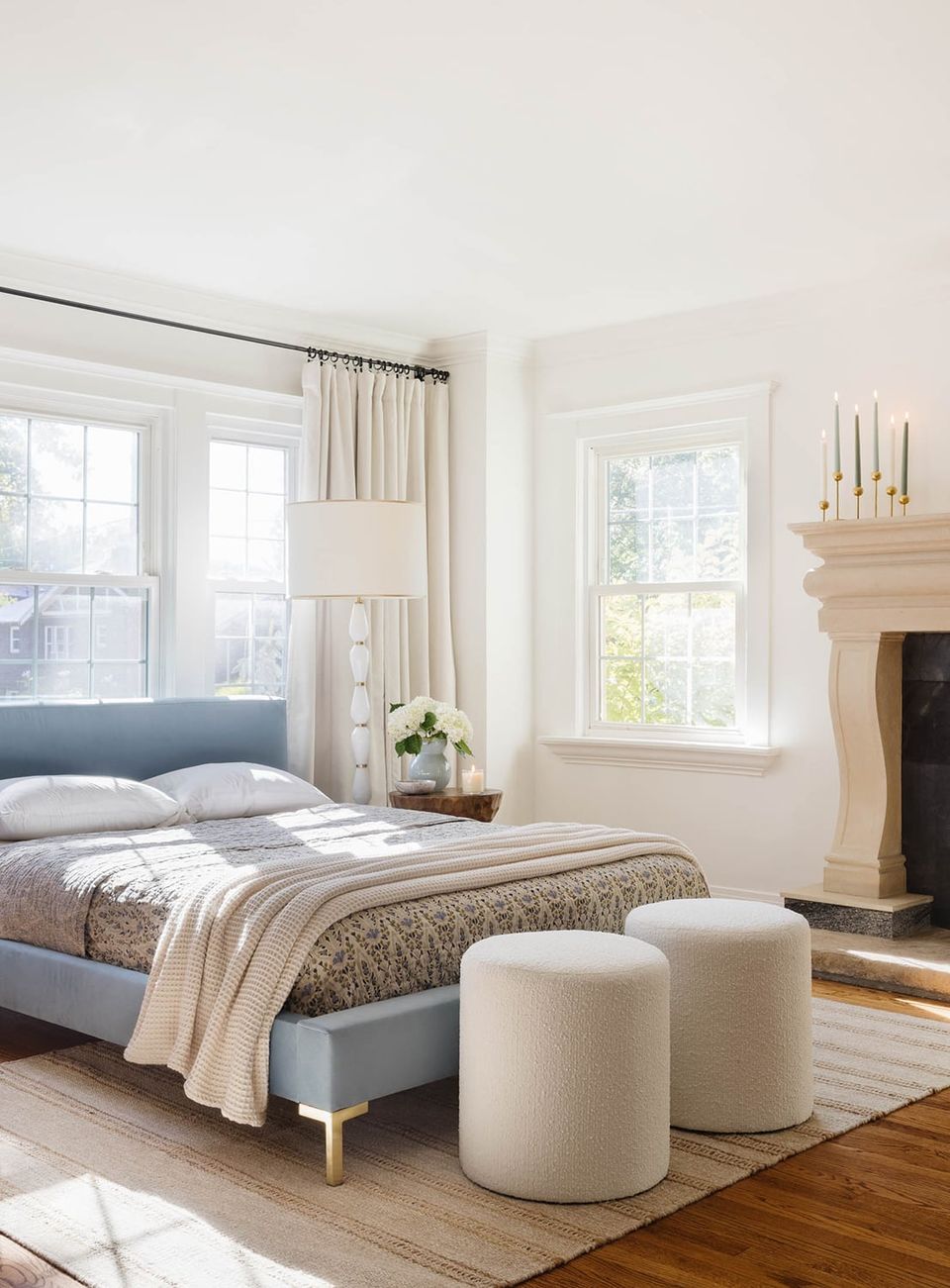 Bright farmhouse bedroom with blue-gray upholstered bed, cream linen drapes, fireplace accent, and sculptural white poufs in warm wood tones.