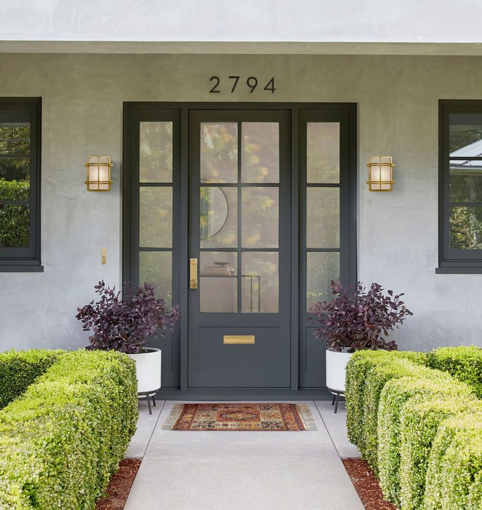 Modern farmhouse entryway with sage-green exterior, black door, symmetrical brass sconces, and manicured boxwood hedges
