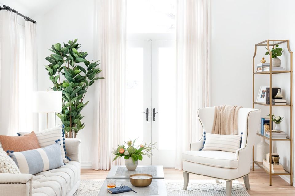 Bright minimalist living room with sheer white curtains, cream armchair, natural wood shelving, and lush green plants creating a calm, airy aesthetic