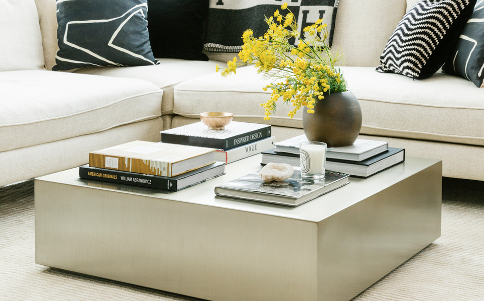 Modern minimalist living room with cream coffee table, stacked design books, yellow flowers, and geometric patterned pillows