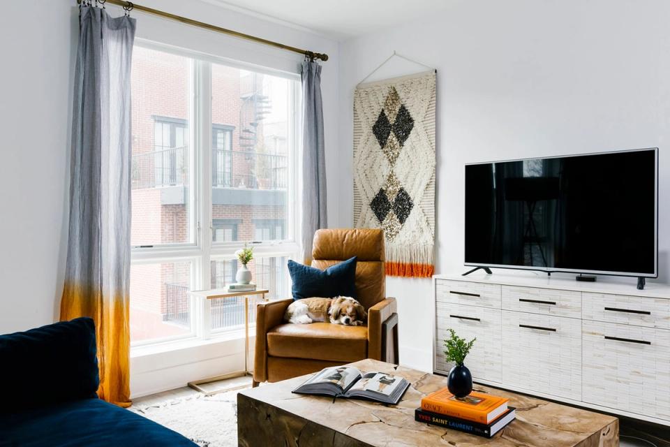 Mid-century modern living room with golden yellow curtains, warm wood chair, white media console, and minimalist decor against soft gray walls