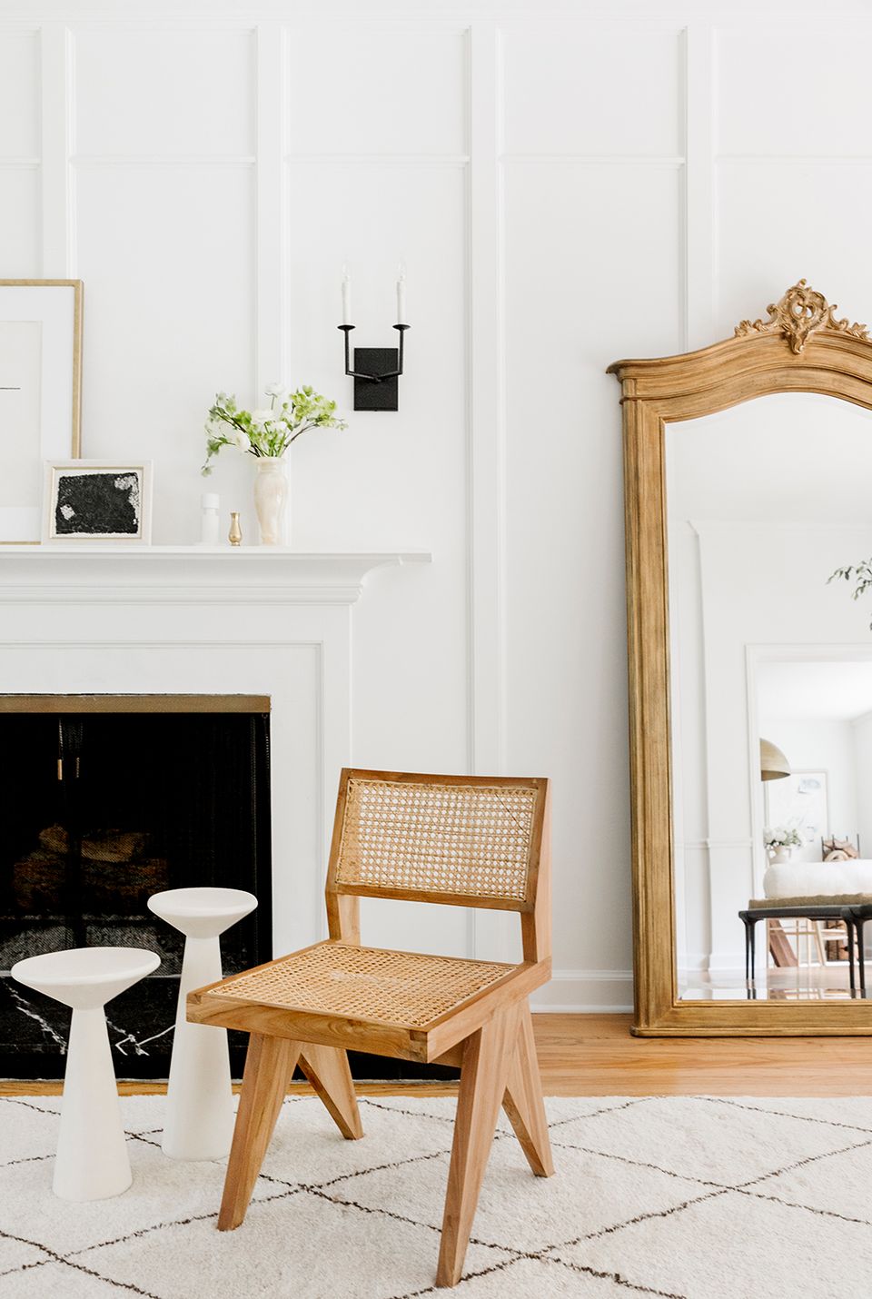 Minimalist room with white walls, wooden cane chair, fireplace, and gold-framed mirror