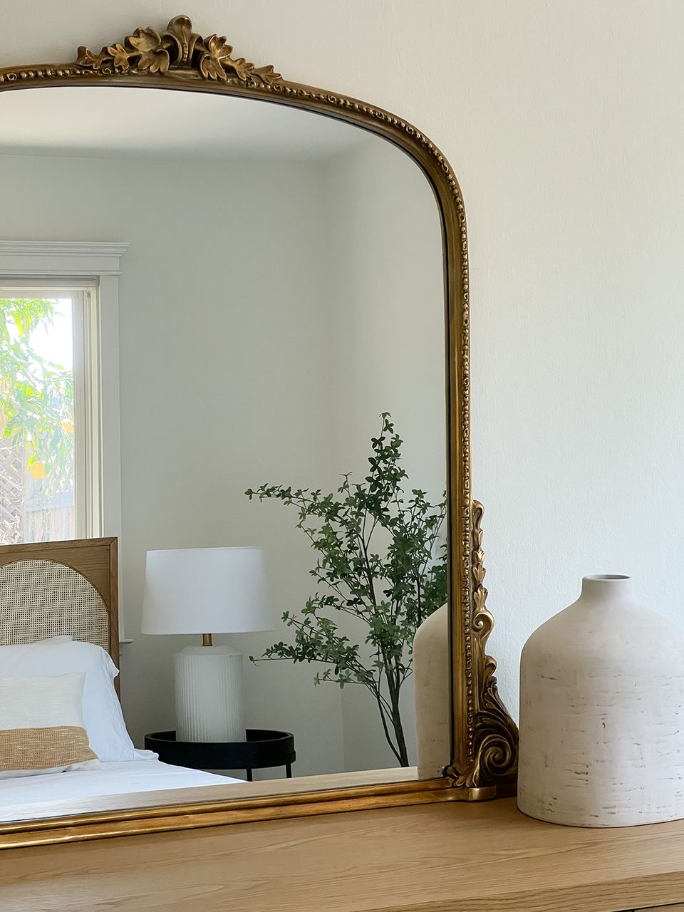 Refined organic modern bedroom detail with statement gold-framed mirror, white linens, and botanical décor
