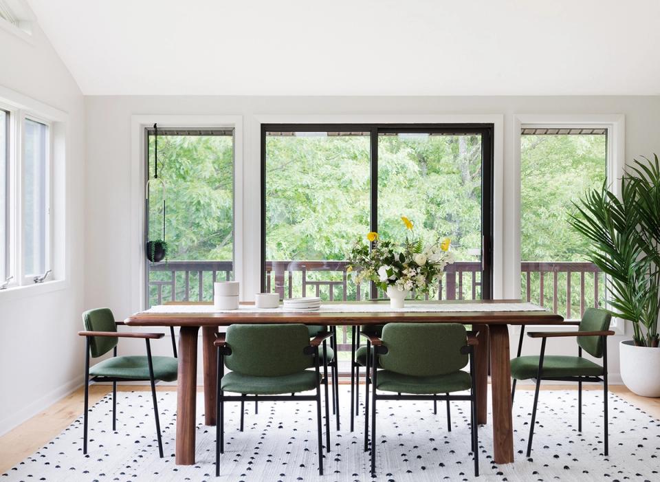 Bright dining room with mid-century modern green chairs, natural wood table, and nature views from large black-framed windows