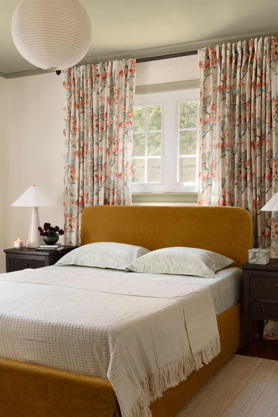 Traditional bedroom with mustard velvet headboard, floral curtains, and soft neutral bedding in warm, inviting setting