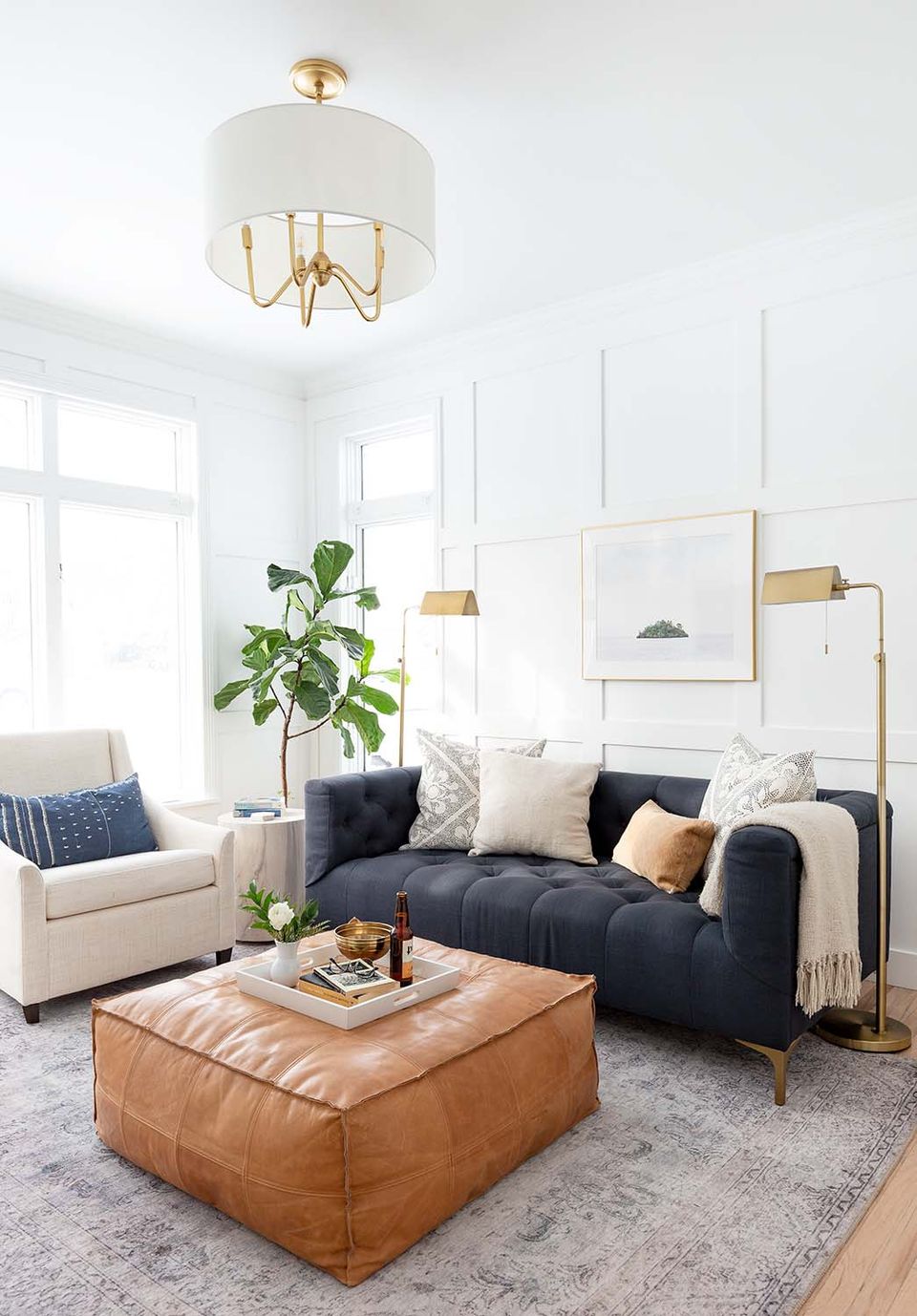 Balanced contemporary lounge with navy sofa, cream accent chair, cognac leather pouf, and brass pendant lighting in soft neutral tones