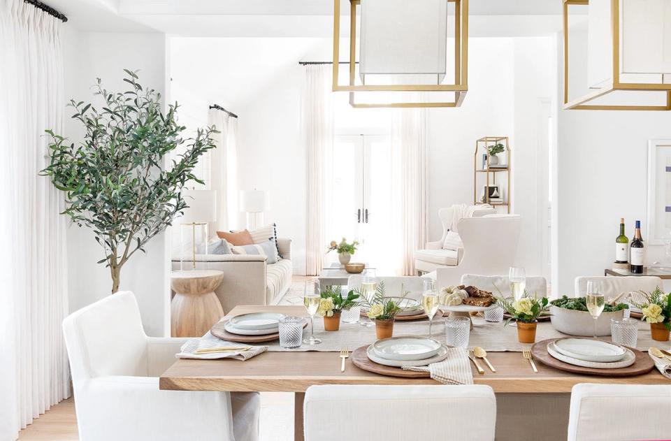 Bright, airy modern dining with natural wood table, white chairs, brass fixtures, and abundant greenery