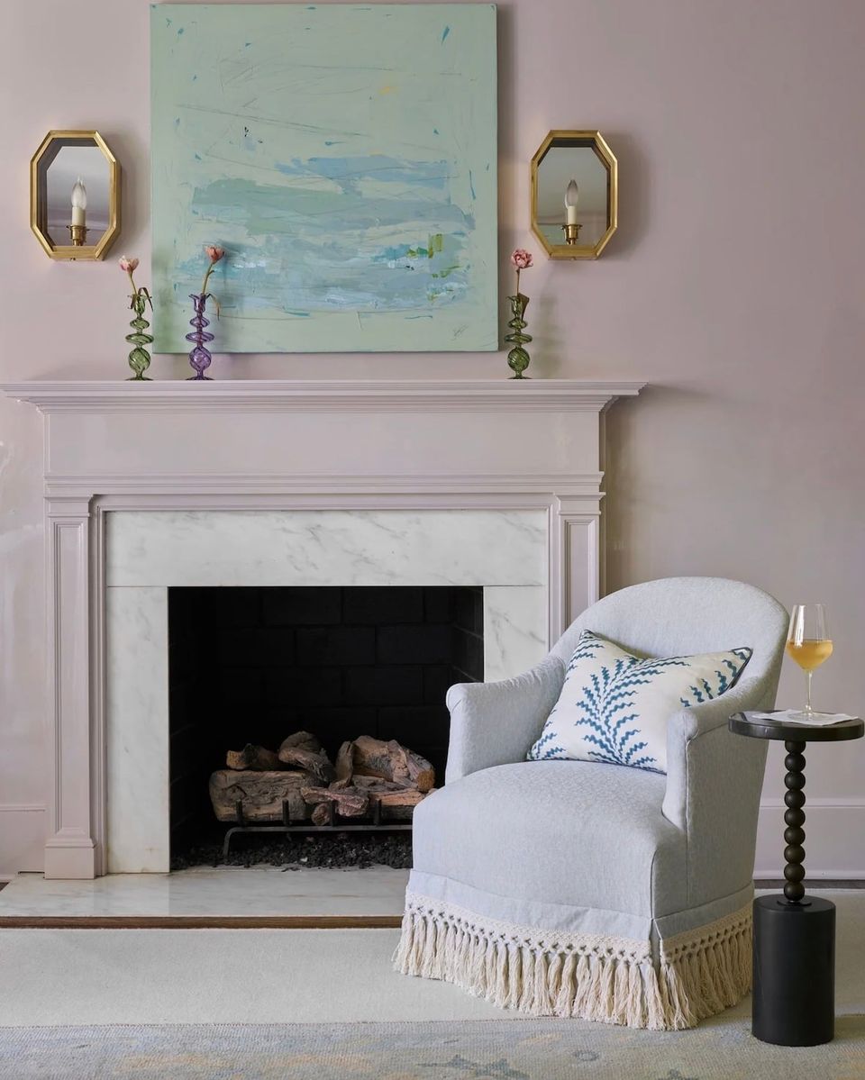 Soft mauve fireplace wall with pale blue abstract art, brass sconces, and cream upholstered chair creating serene, elegant atmosphere