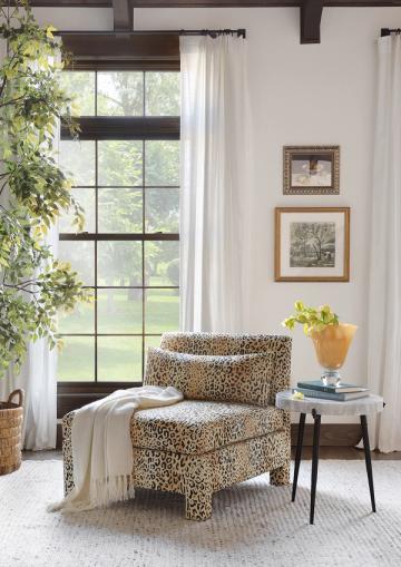 Contemporary farmhouse corner with leopard-print chair, natural wood side table, and large windows with dark beams.