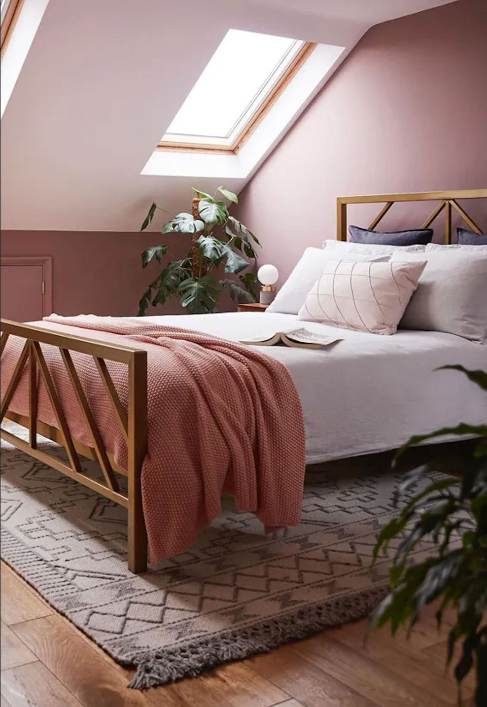 Soft mauve bedroom with natural skylight, mid-century wood bed frame, layered textures, and potted greenery creating a serene, botanical retreat.