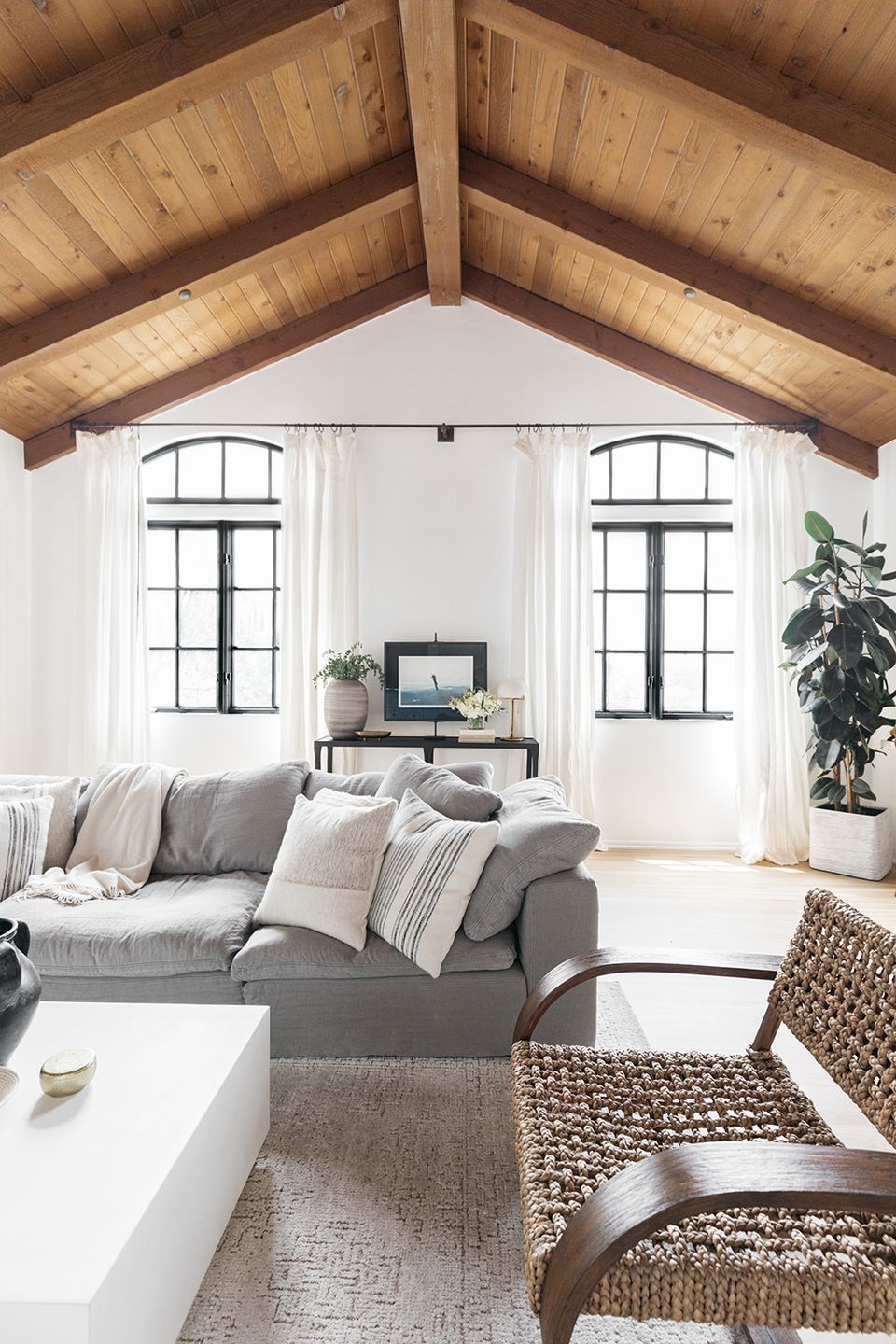 Bright farmhouse living room with exposed wooden beams, gray sofa, natural textures, and abundant natural light from arched windows