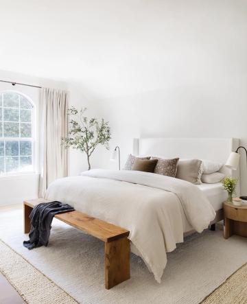 Minimalist farmhouse bedroom with soft cream linens, natural wood bench, and diffused window light for serene simplicity.