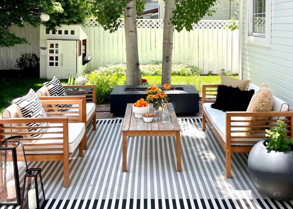 Modern coastal patio with striped outdoor rug, wood furniture, black cushions, and bright florals under shade trees