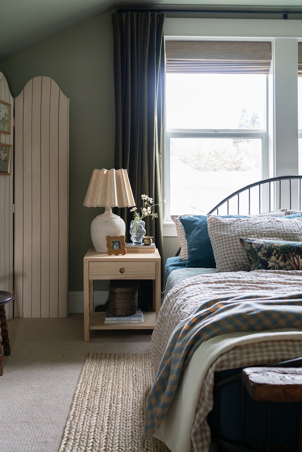 Cozy traditional guest room with sage green walls, blue plaid bedding, natural wood nightstand, and soft lamplight.
