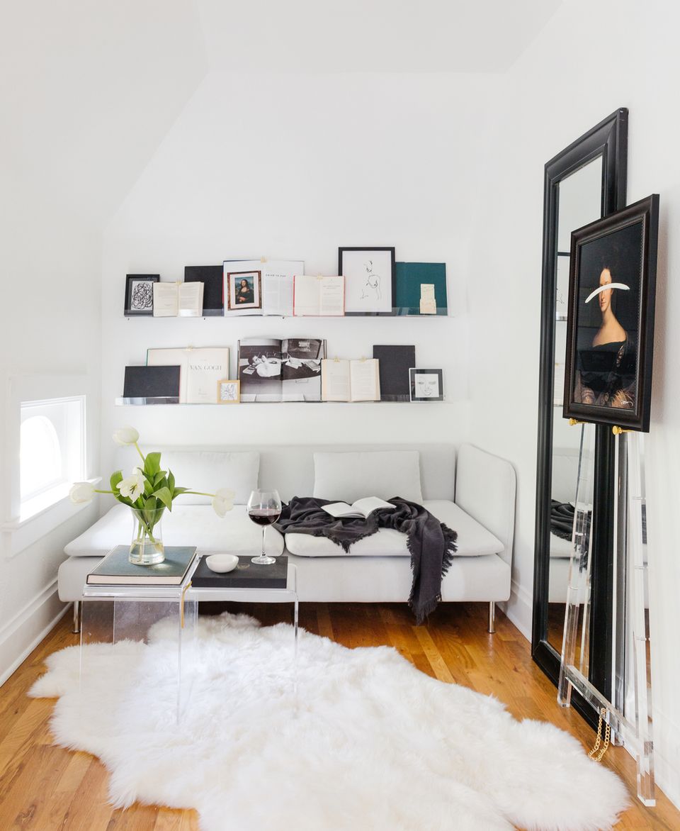 Minimalist reading nook with white sofa, floating shelves displaying curated objects, black-framed mirror, and warm wood flooring accented by white fur rug.