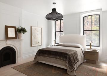 Farmhouse bedroom with neutral palette, white upholstered headboard, dark plaid throw, and fireplace accent creating serene countryside charm