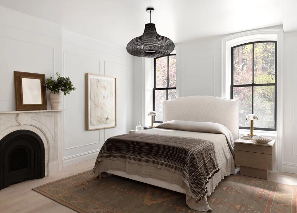 Farmhouse bedroom with neutral palette, white upholstered headboard, dark plaid throw, and fireplace accent creating serene countryside charm