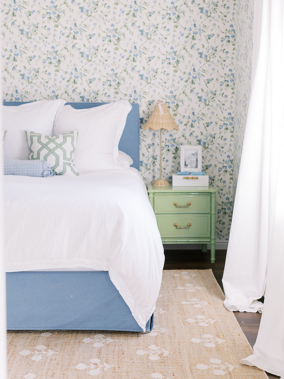 Airy preppy bedroom featuring floral wallpaper, blue bed frame, white linens, and sage green nightstand