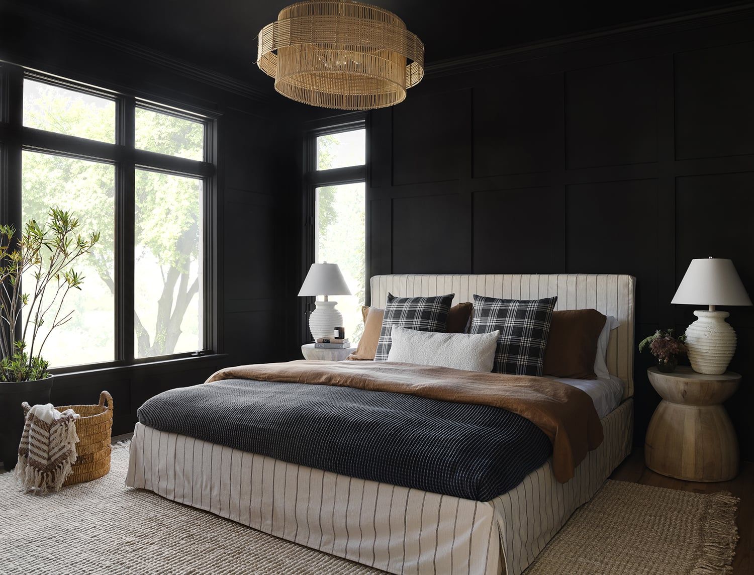 Dramatic black bedroom with natural light, cream striped upholstered bed, warm wood accents, and woven pendant fixture