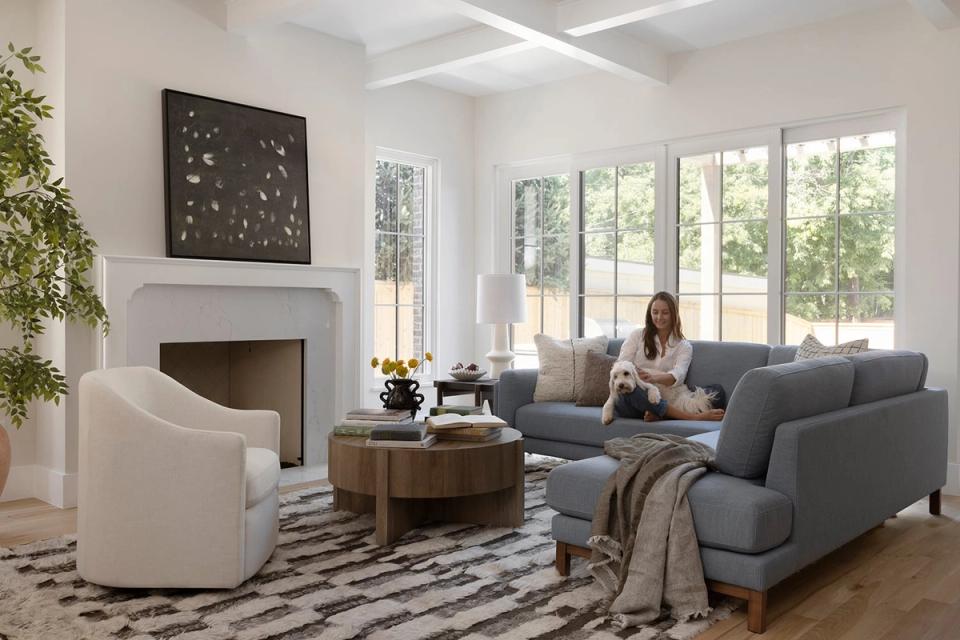 Modern transitional living room with gray sectional, white armchair, geometric patterned rug, and fireplace with natural light.