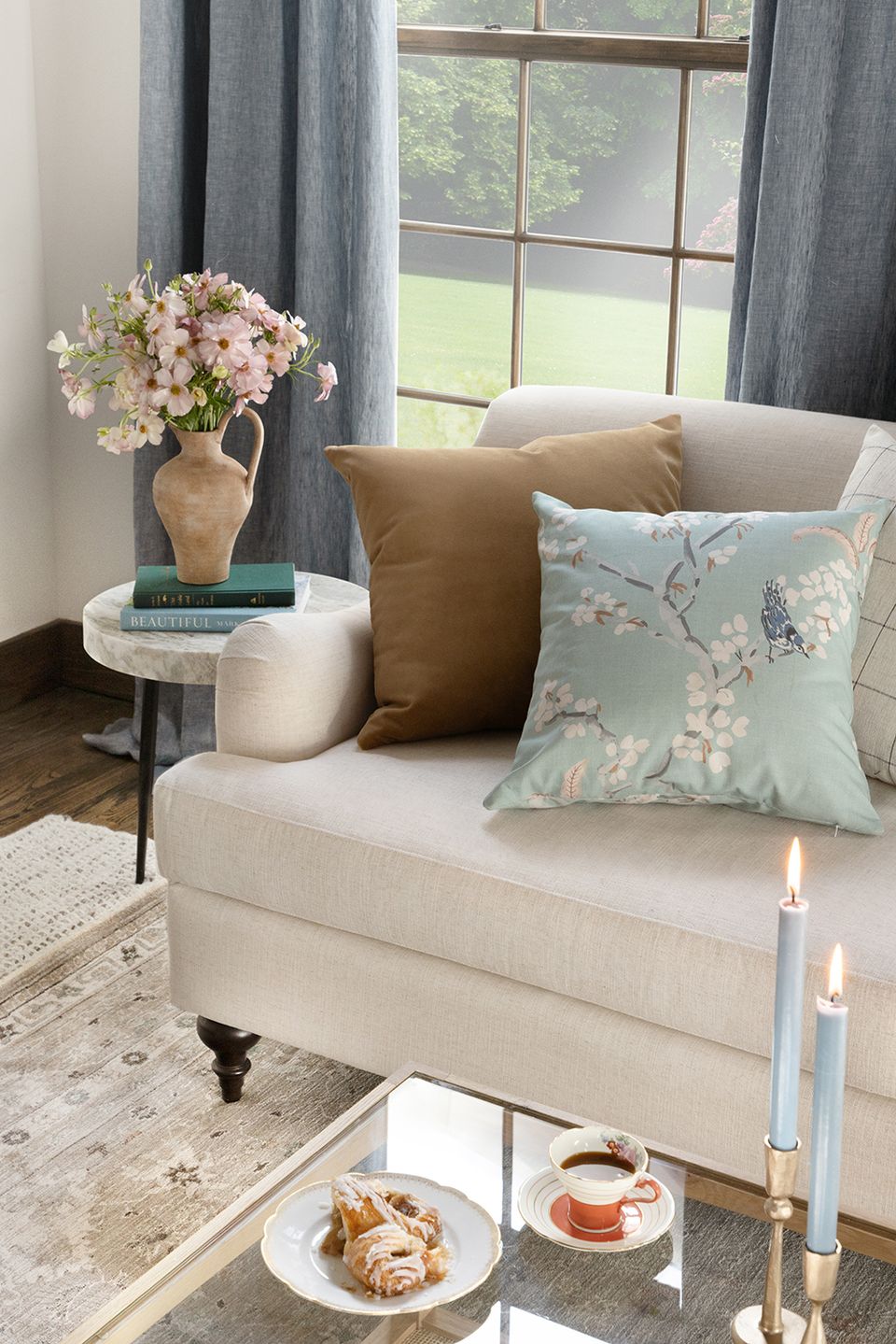 Serene traditional living room with cream upholstered seating, sage floral pillows, dusty blue linen curtains, and warm terracotta pottery accents.