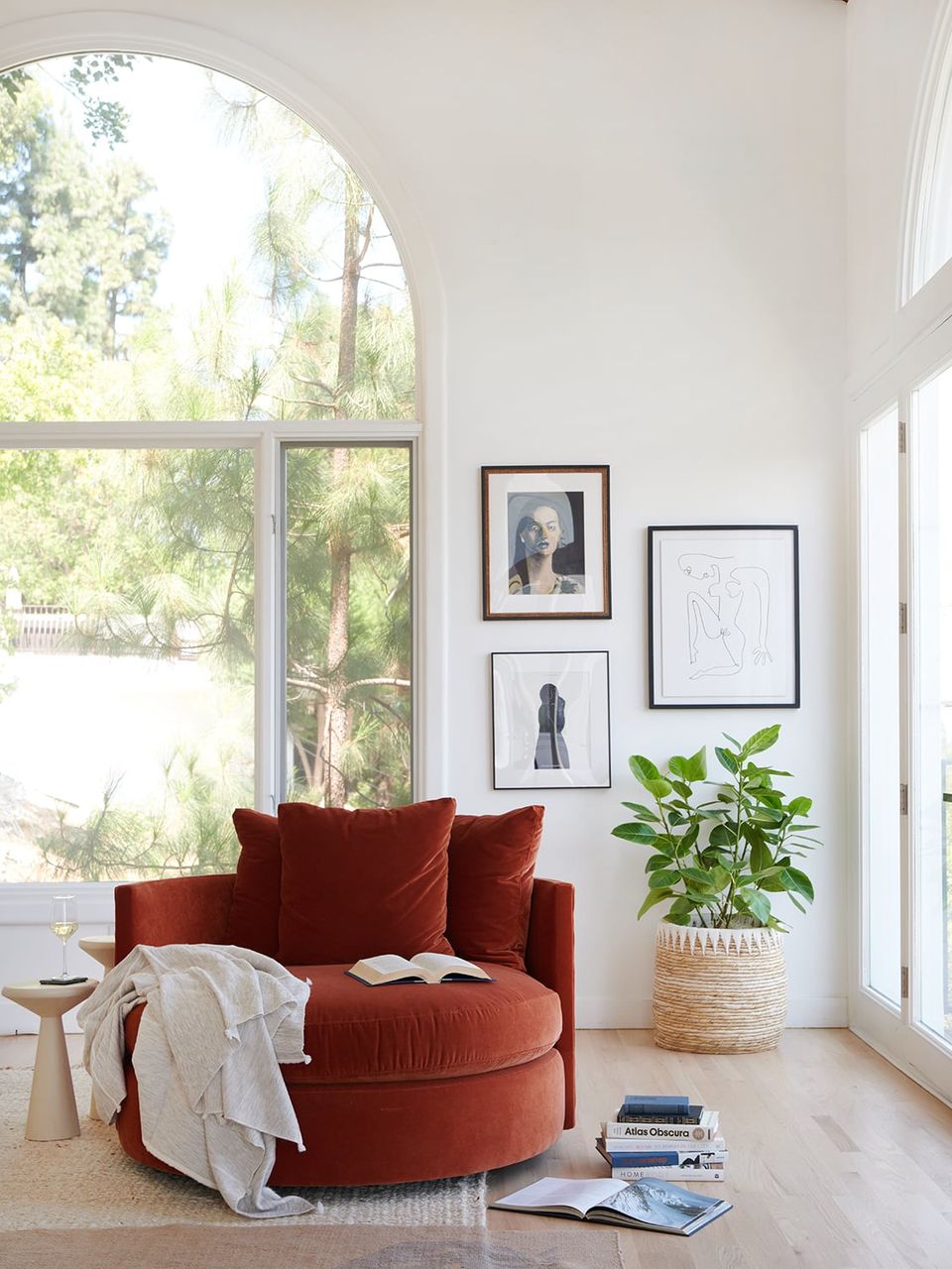 Minimalist salon with burgundy round chair, scattered framed prints, potted plant, and arched window creating serene modern elegance.
