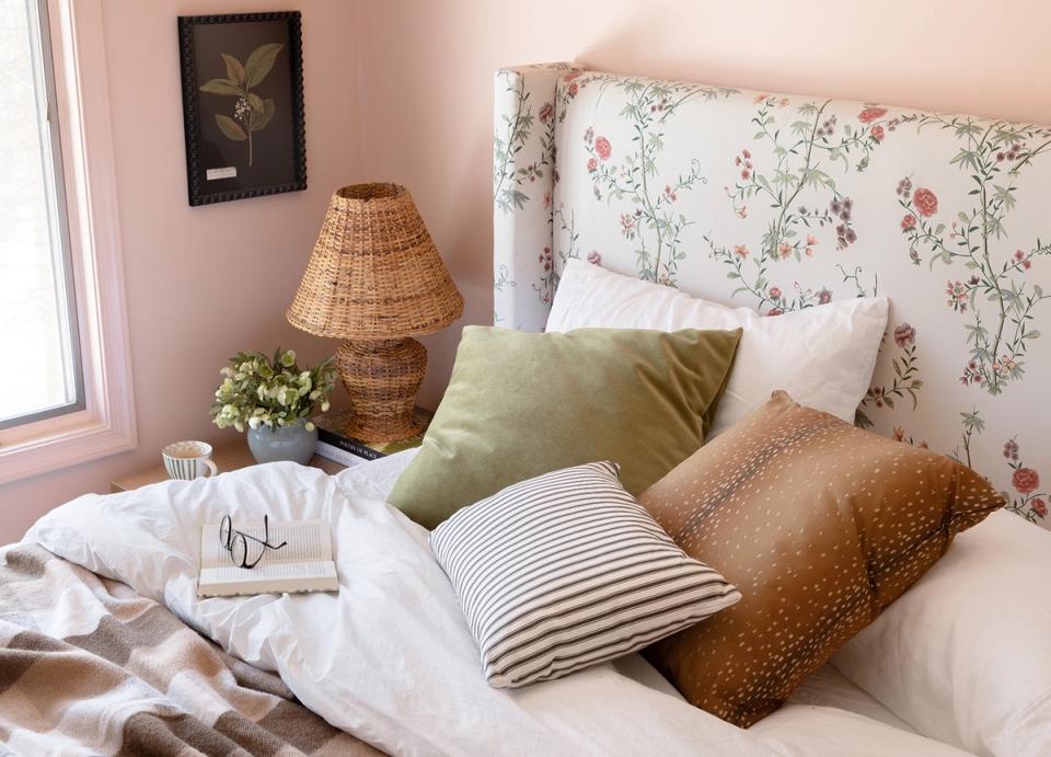 Romantic bedroom in soft pink tones with floral headboard, layered textured pillows, and rattan lamp.