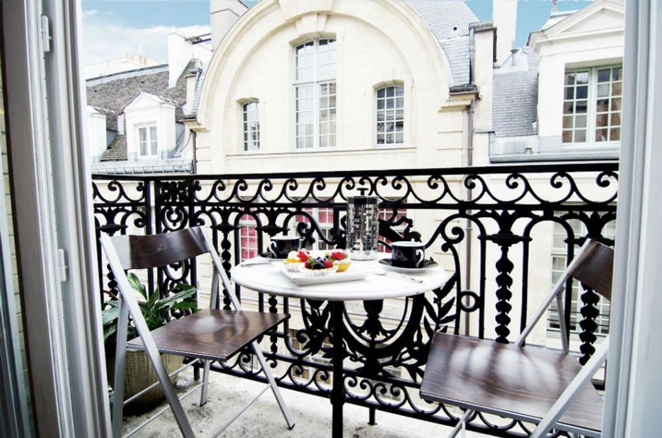 Parisian-style balcony with ornate black iron railings, round white table, classic bistro seating, and elegant architectural details overlooking rooftops.